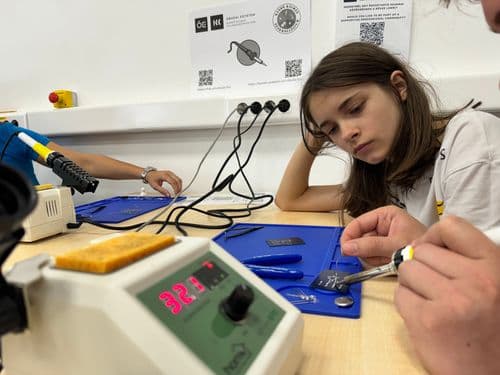 Egy tanuló lány a forrasztást gyakorolja, előtérban a forrasztó készülék, amin a tanár keze látszik még, a háttérben a kislány könyökölve nézi