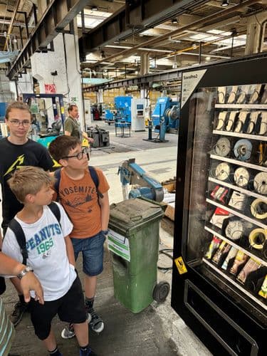 PO tábor 2024 - érdekes automata a BKV telephelyén: három tanuló nézi a csiszolókorong, ragasztó és egyéb munkaeszközöket kínáló automatát, amit a dolgozók a kártyájukkal működtetnek