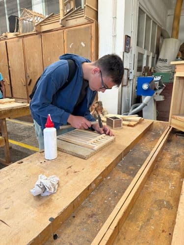 PO tábor 2024 - tanuló kenyértartót eszkabál az asztalosműhelyben, a szemüveges fiú éppen kalapáccsal szögeli össze a vékony léceket