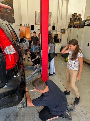 PO tábor 2024 - Tanulók autókereket cserélnek a tanműhelyben. Egy fiú próbálkozik egy autó hátsó kerekénél a kerékkulccsal