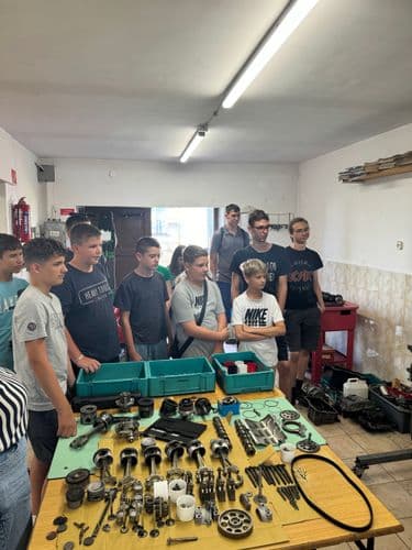 PO tábor 2024- a tanulók az autószerelő műhelyben állnak, előtérben egy asztal, gondosan rendben kirakott alkatrészekkel: ékszíjak, csapágyak, dugattyúk , tömítések vannak rajta