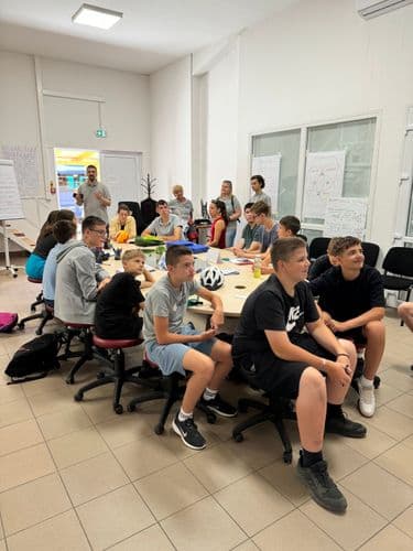 PO tábor 2024-Tanulók csoportja ül a DKA aulájában az ovális asztal körül és az oktatóra figyel