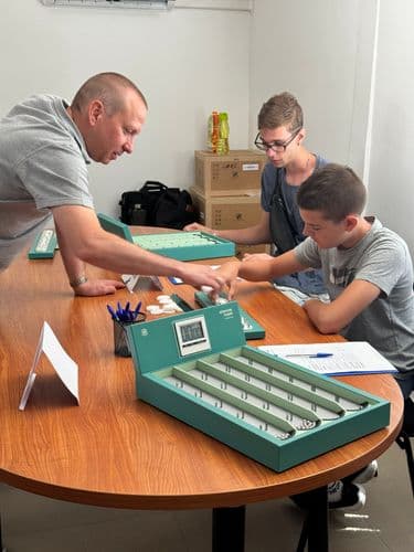 PO tábor 2024 - a logisztikai játék nagy doboza, egy oktató magyarázzal a játékszabályokat két fiúnak, akik egy ovális asztalnál ülnek