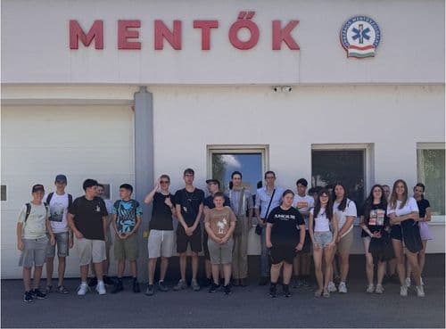 A Váci Mentők bejáratánál állnak a résztvevő gyerekek, újabb csoportkép. Felettük a mentők logója és a felirat, a garázsajtó előtt